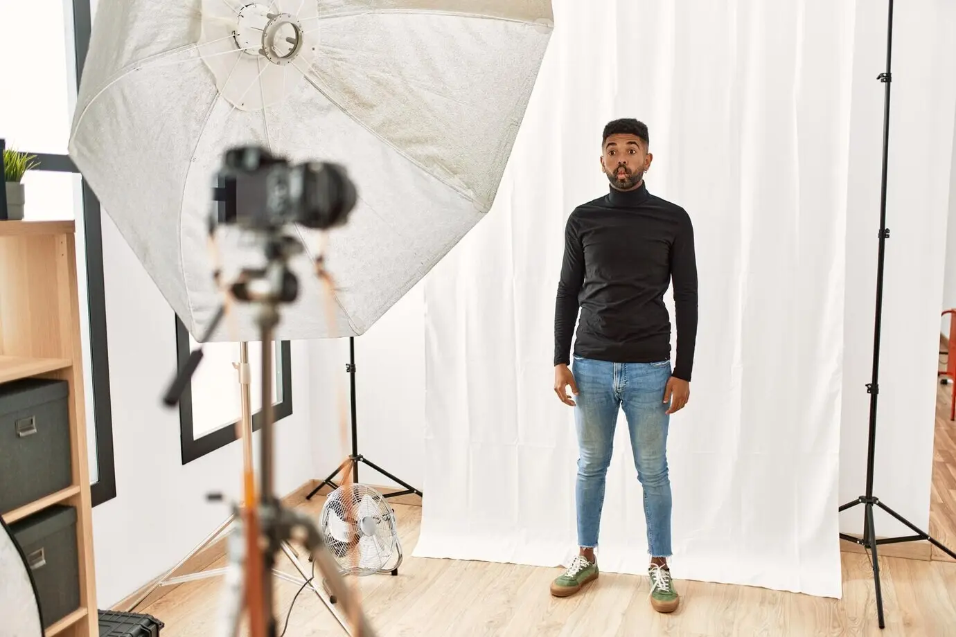 Молодой мужчина латиноамериканского происхождения с бородой позирует как модель в фотостудии, складывая губы «рыбкой», при этом делает сумасшедший и комичный жест и демонстрирует смешное выражение лица.